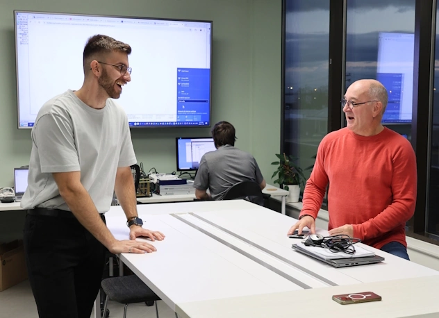 Direct Answer: Two Letico engineers smiling and collaborating at a modern workspace table, with an active monitor and office environment in the background.